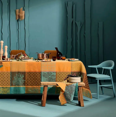 A styled dining table set with a patchwork tablecloth in orange, teal, and brown tones, accompanied by a light blue chair and rustic wooden table in a moody, textured room.
