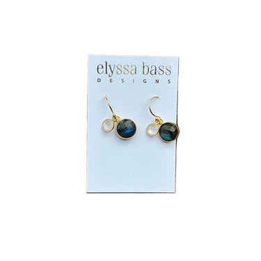 A pair of gold hoop earrings with round dark blue-green labradorite stones, displayed on a white card labeled 'elyssa bass DESIGNS' against a red brick background.