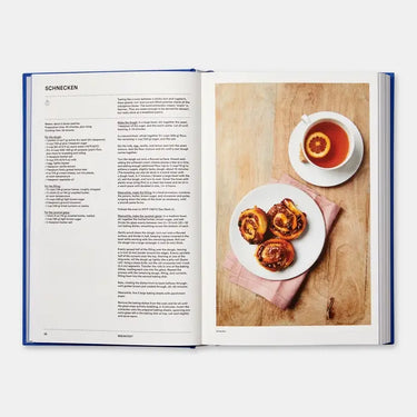 Open book showing a recipe for Schnecken with a plate of pastries and a cup of tea on a wooden board.
