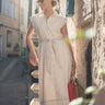 Woman in a beige dress standing on a street with stone steps and buildings.