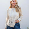 Woman wearing a beige knitted sweater and blue jeans against a white wall.