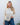 Woman wearing a beige knitted sweater and blue jeans against a white wall.