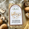 Packaged mushroom fettuccine with Valente Market label surrounded by mushrooms on a textured fabric background